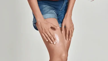 A person in denim shorts applies hydrocortisone cream to their thigh, conveying a sense of self-care and attention to skincare on a neutral background.